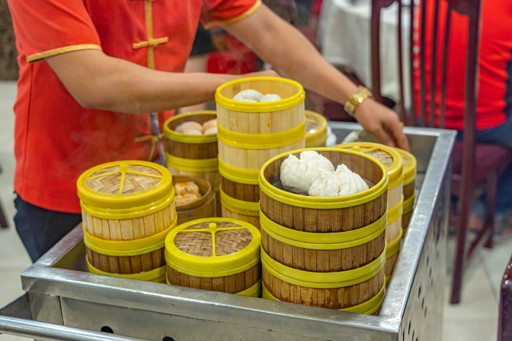 A server in a red uniform arranges stacked bamboo steamer baskets filled with dim sum on a metal cart.