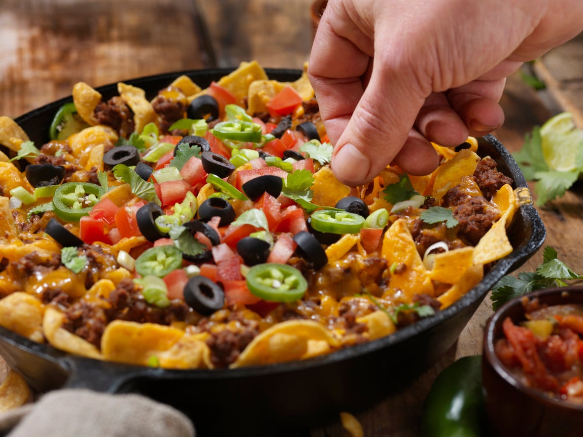 A hand picks up a nacho topped with ground beef, cheese, black olives, jalapeños, and tomatoes from a cast-iron skillet.