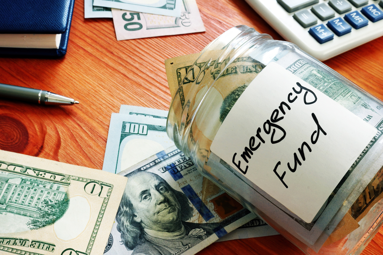 A glass jar labeled “Emergency Fund” filled with US dollar bills, surrounded by loose cash, a pen, a notebook, and a calculator on a wooden surface.