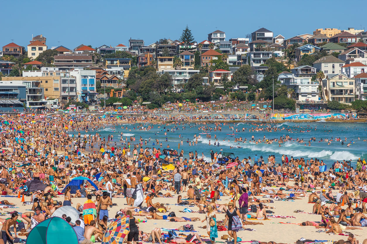 A crowded Bondi Beach in Australia filled with sunbathers and swimmers, with hillside houses overlooking the ocean, perfect for informing trip planning decisions.