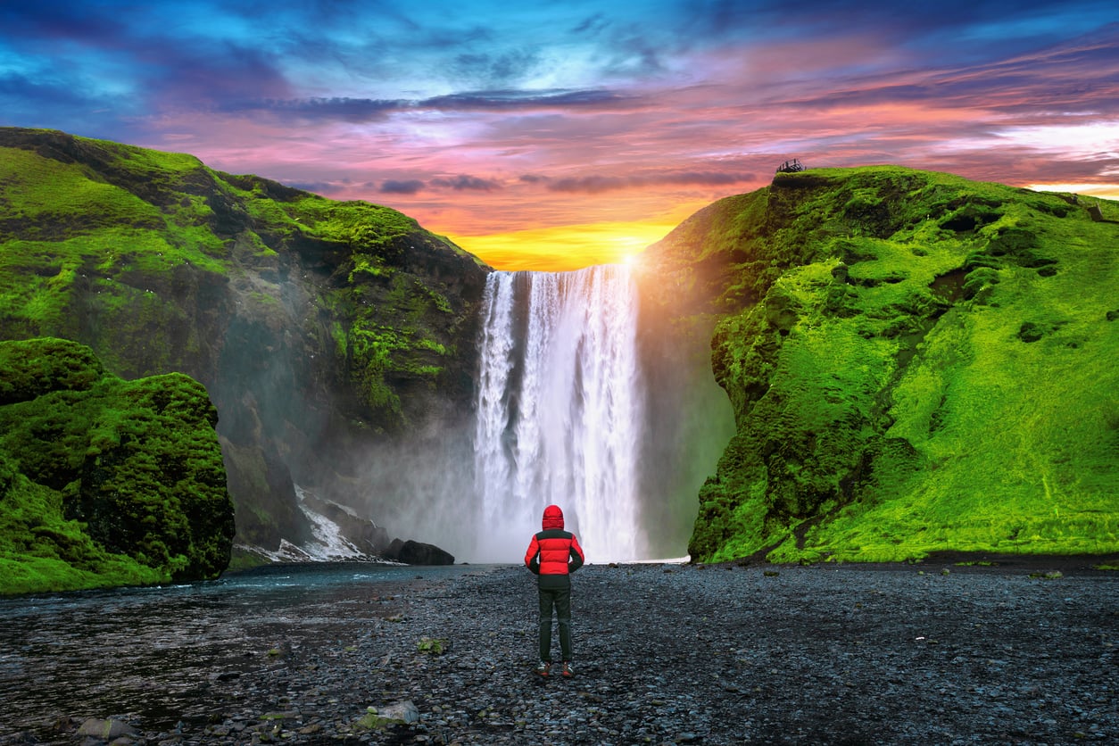 A person in a red jacket stands on a dark pebble beach facing Skógafoss waterfall under a vibrant sunset sky.