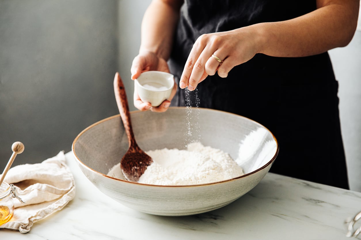 Hands sprinkling salt into a large mixing bowl filled with flour.