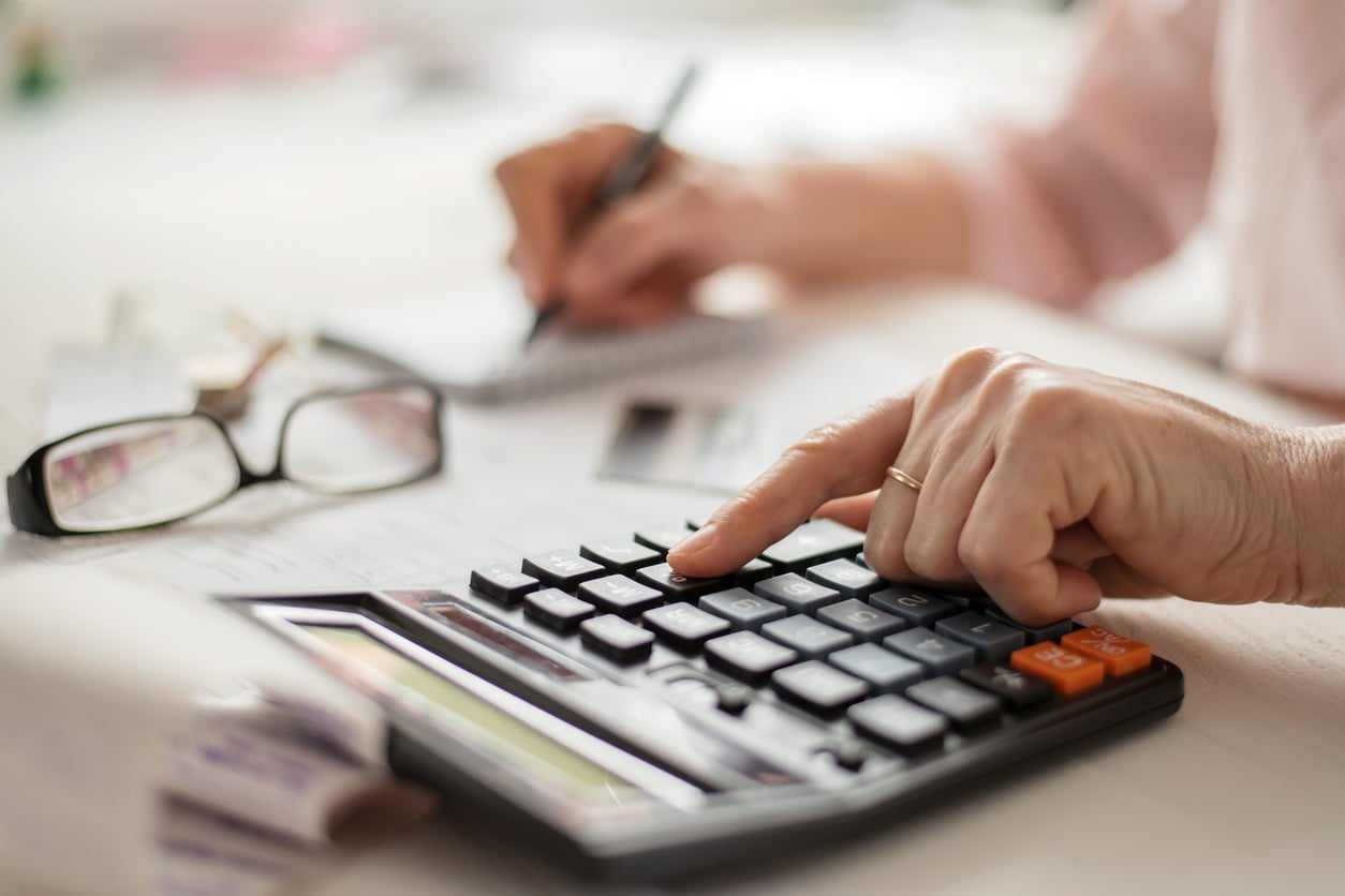 A person uses a calculator while writing in a notepad, with reading glasses on the table nearby.