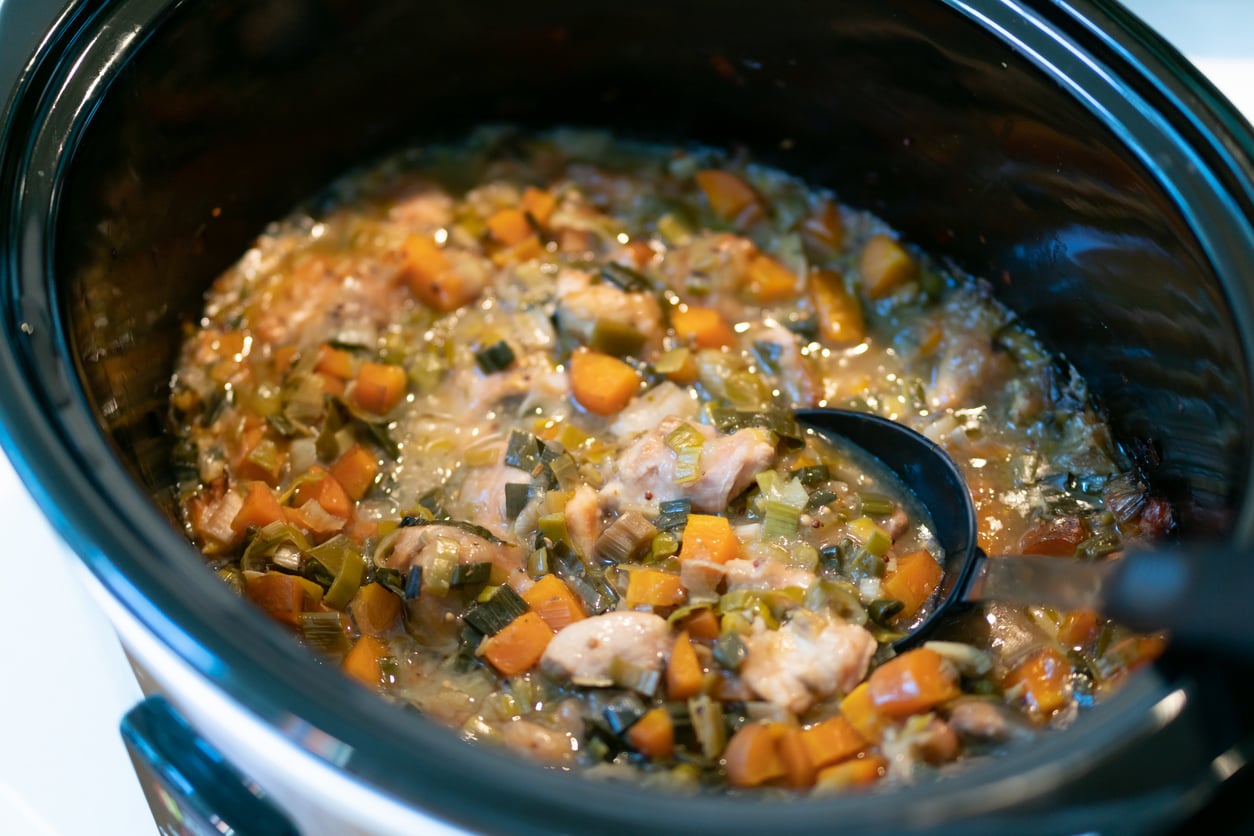 A slow cooker filled with chicken and vegetable stew, with a black ladle submerged in the center.
