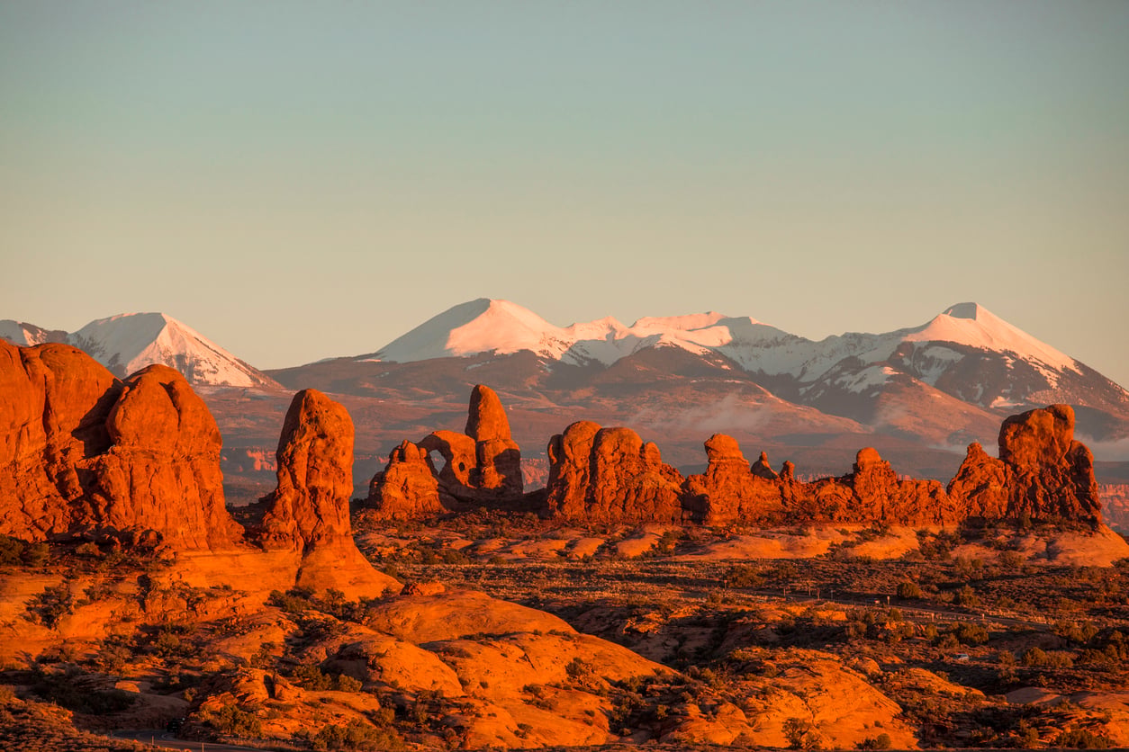 Red rock formations rise from the desert floor with snow-capped mountains in the background, illuminated by warm sunlight—perfect for vacations where the hotel barely matters.