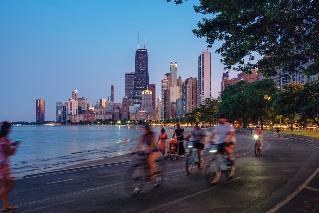 People cycling and walking on a path along a lakefront with the Chicago skyline in the background at dusk.