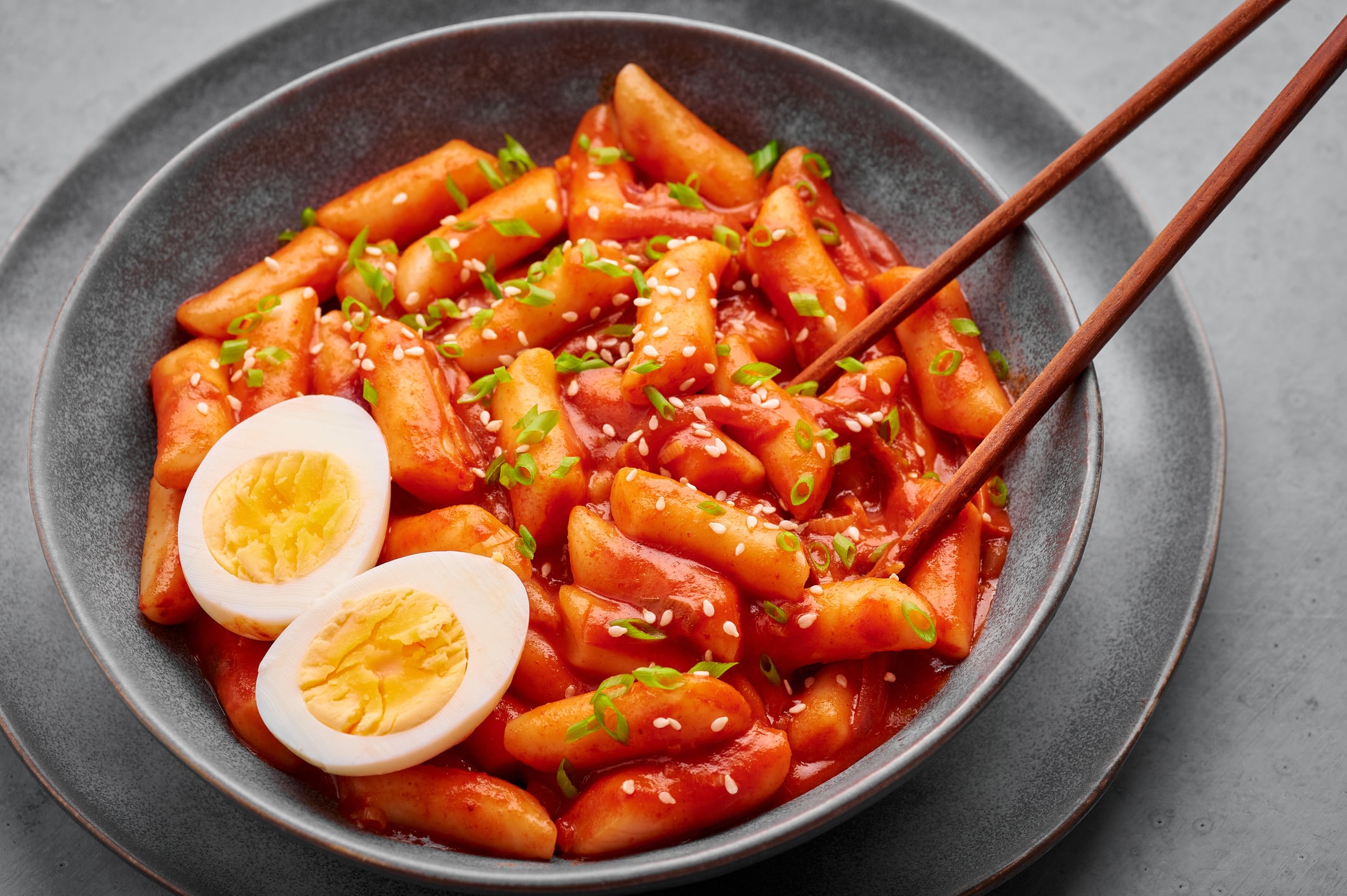 A bowl of tteokbokki topped with sliced hard-boiled egg, sesame seeds, and chopped scallions, with a pair of wooden chopsticks resting inside.