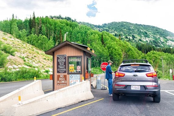 A person stands outside a wooden fee booth at the entrance to a forest area, talking to the driver of a gray SUV stopped at a red sign.