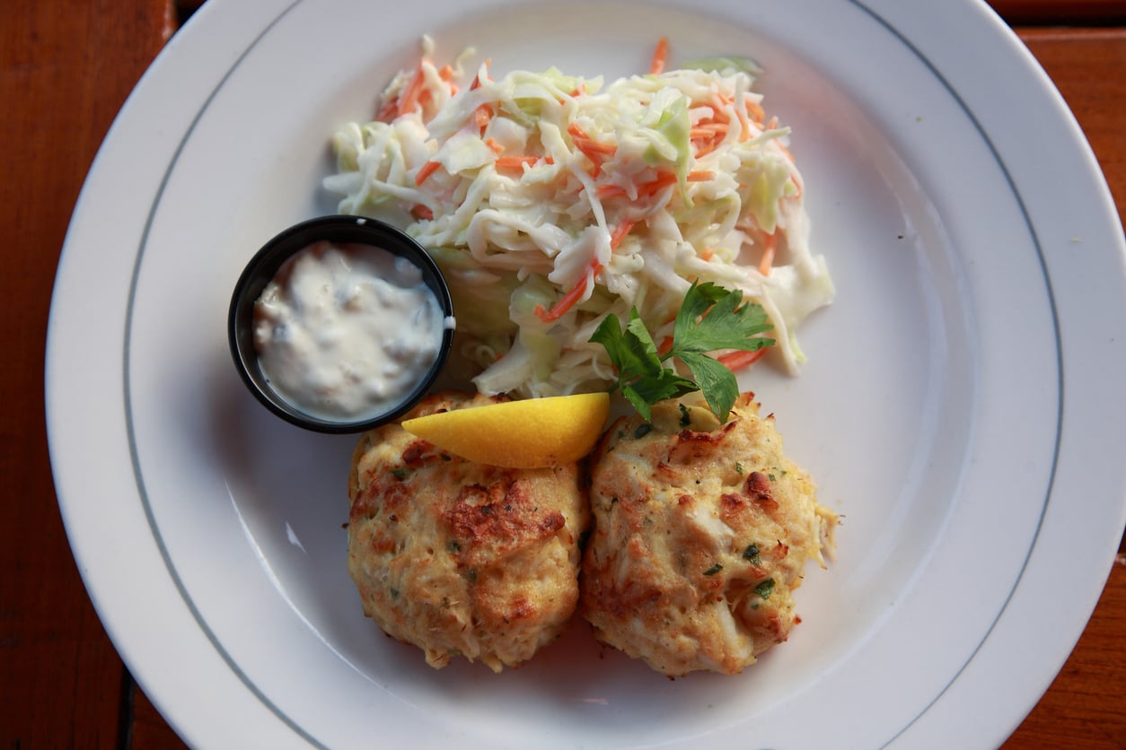 Two crab cakes on a plate with a side of coleslaw, a lemon wedge, and a small cup of tartar sauce.