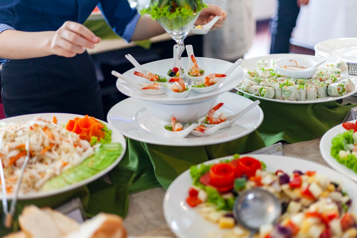 A buffet table displaying various appetizers, including shrimp on spoons, spring rolls, and colorful salads, as a person reaches for a dish.
