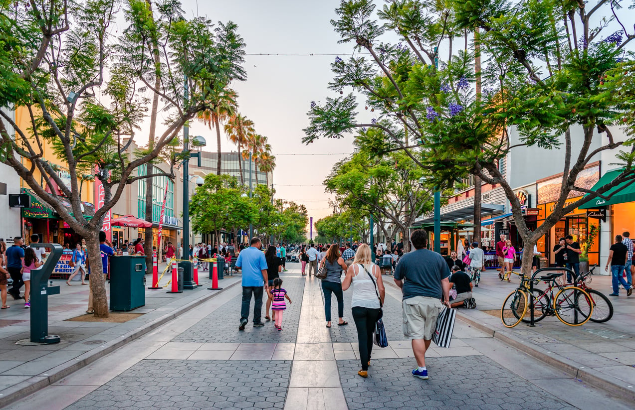 Pedestrians walk along a paved outdoor shopping promenade lined with trees, storefronts, and outdoor seating.