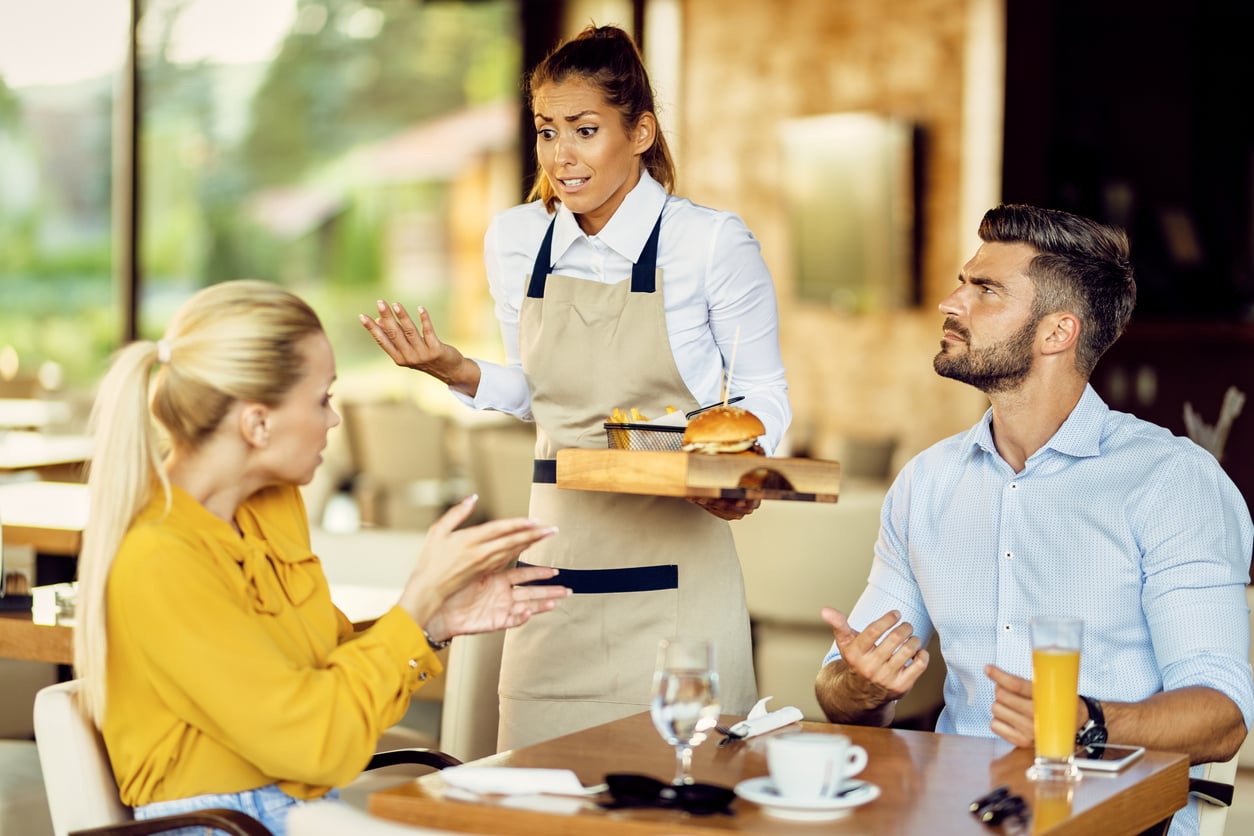 A waitress holding a tray with a burger and fries looks stressed while talking to a man and a woman sitting at a restaurant table.