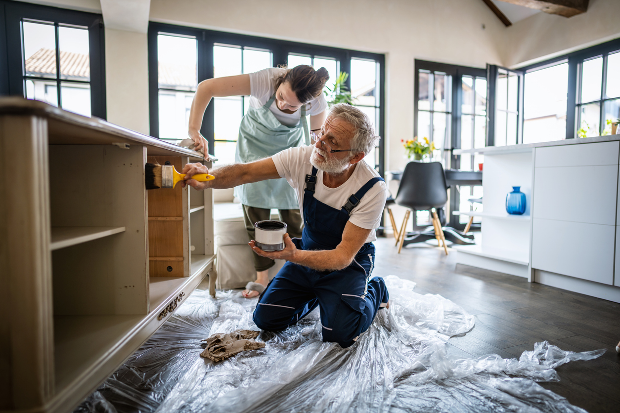A woman and a man in overalls work together to paint a wooden cabinet in a bright room.