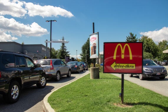 Cars line up in a McDonald’s drive-thru, with a prominent red McDonald’s sign pointing toward the lane.