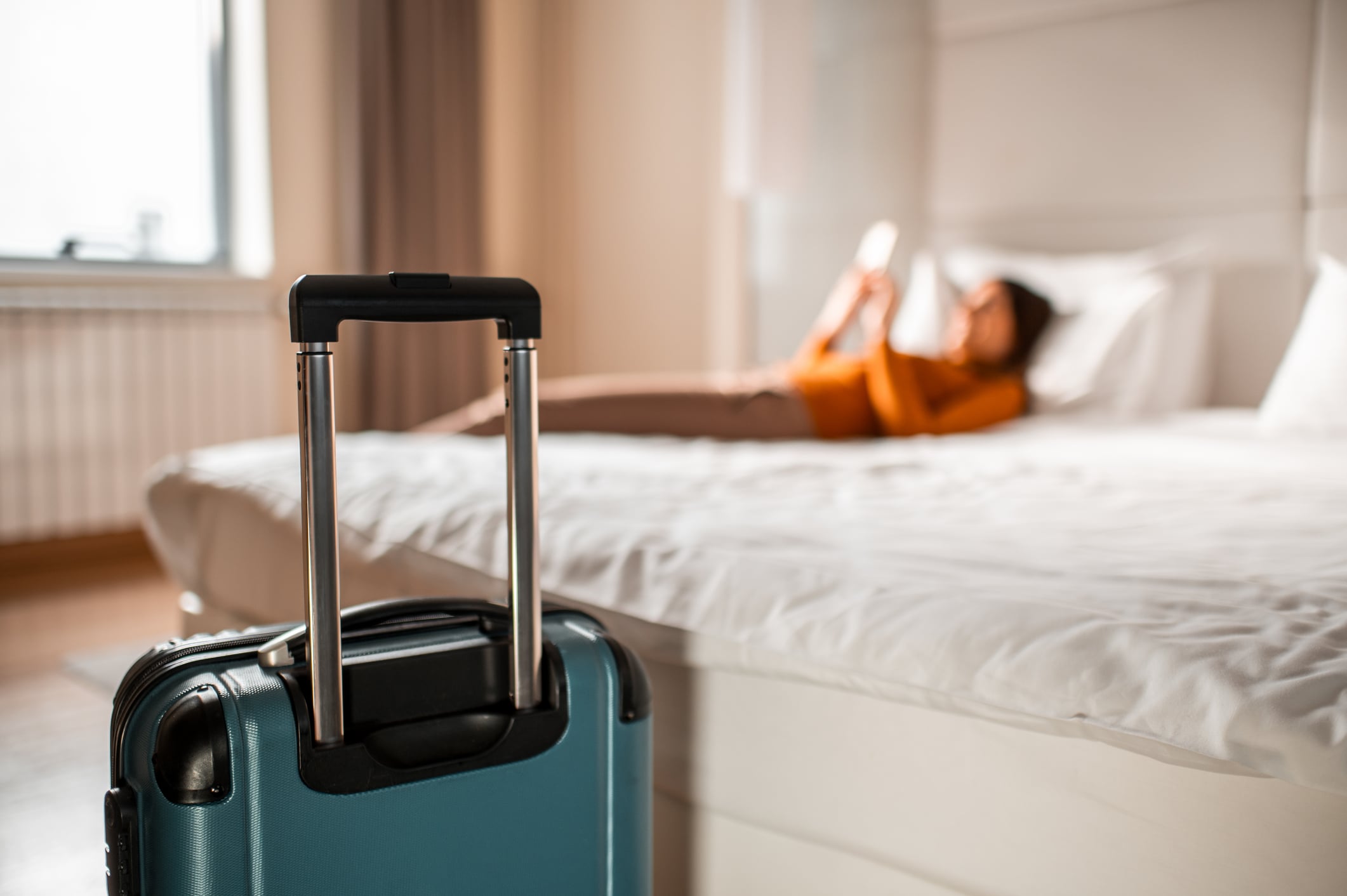 A teal carry-on suitcase stands in the foreground, with a person lying on a hotel bed in the blurred background.