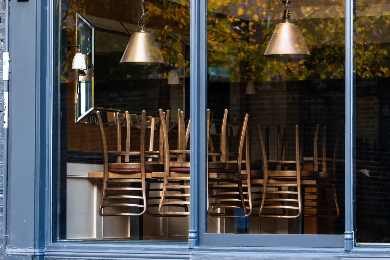 Wooden dining chairs turned upside down on tables inside a restaurant viewed through a window.