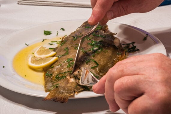 Hands using a knife and fork to cut a whole cooked fish garnished with herbs and lemon slices on a plate.