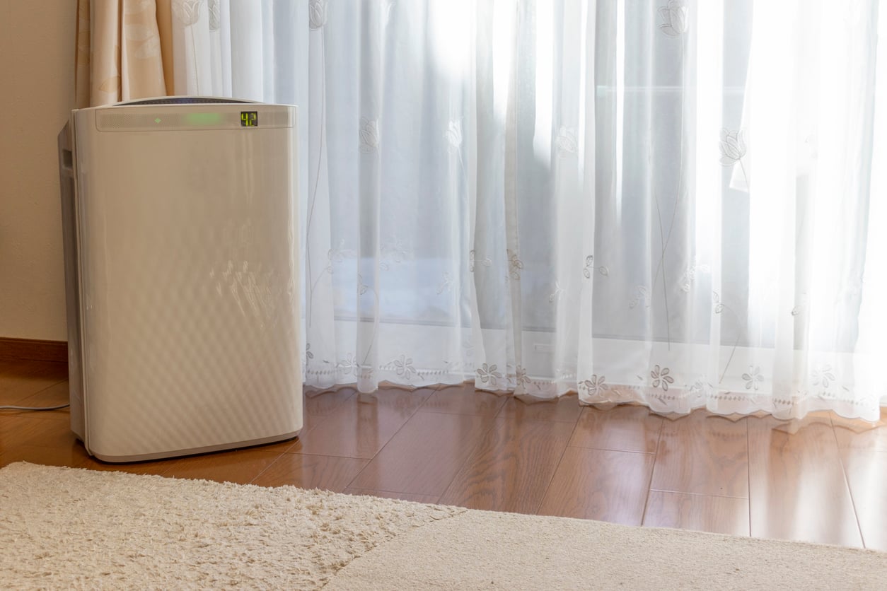 White air purifier with a digital display showing the number 42, placed on a wooden floor in front of a window with sheer curtains.
