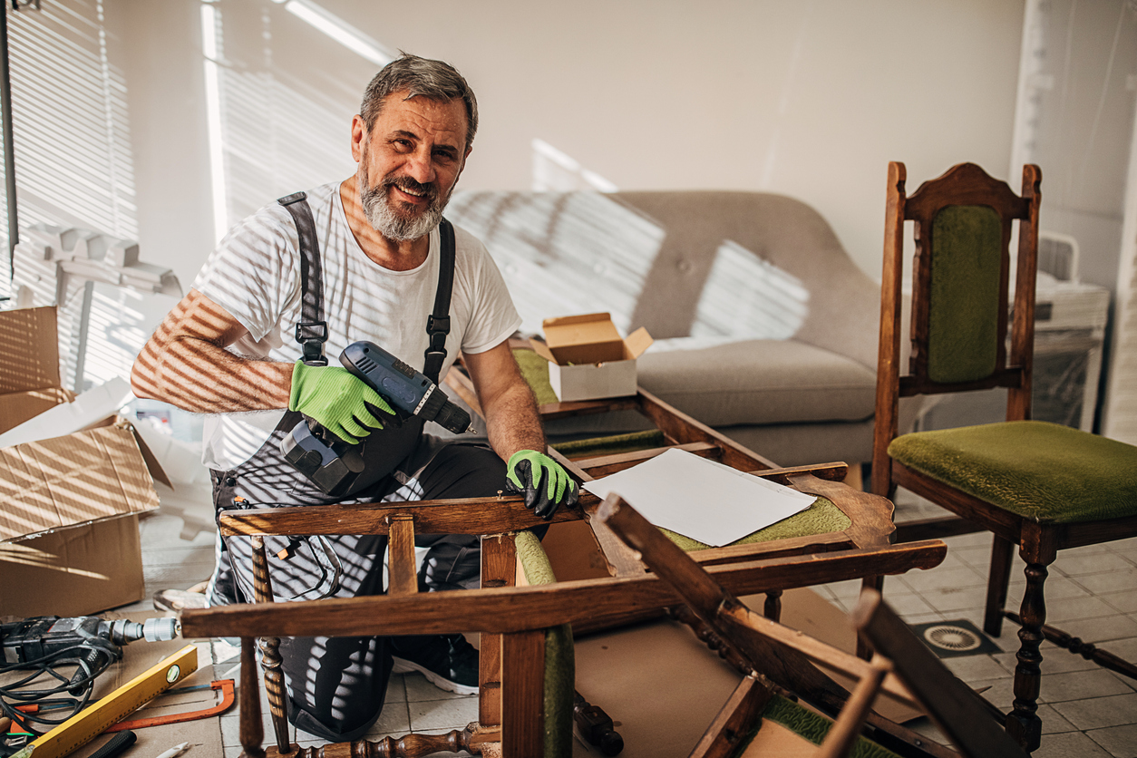 A man wearing work clothes and green gloves uses a power drill to repair a wooden chair.