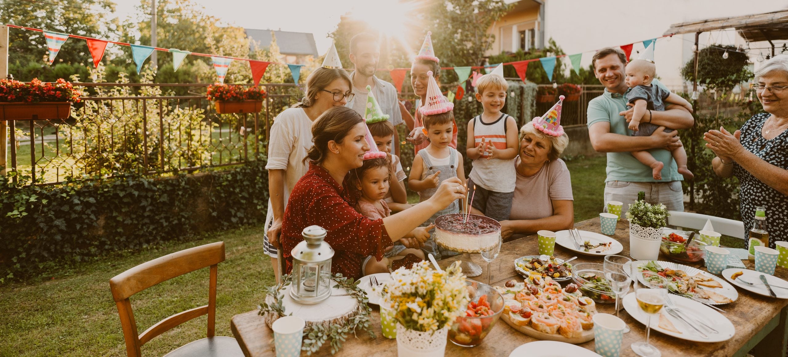 A family gathers around a woman lighting candles on a birthday cheesecake at an outdoor table decorated with party bunting.
