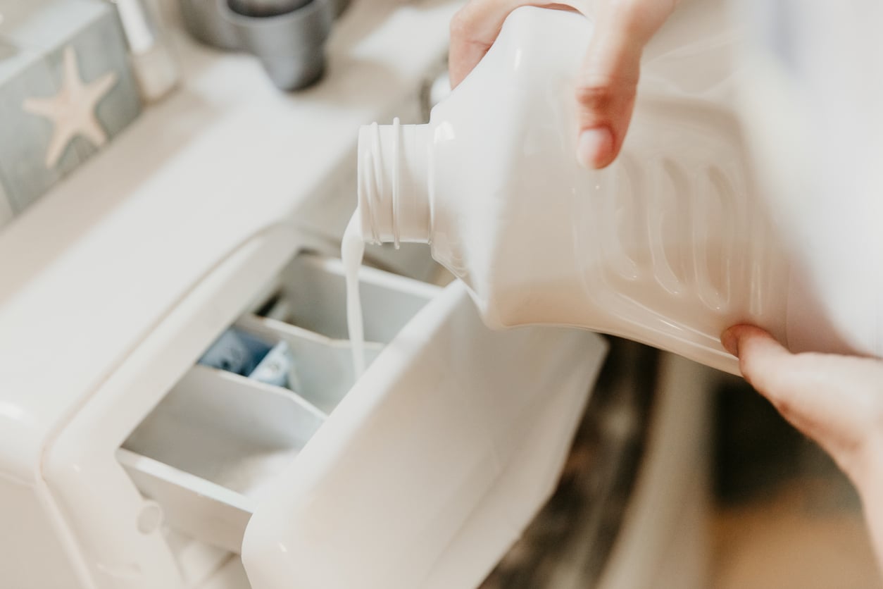 Hands pouring liquid laundry detergent into the dispenser tray of a washing machine.