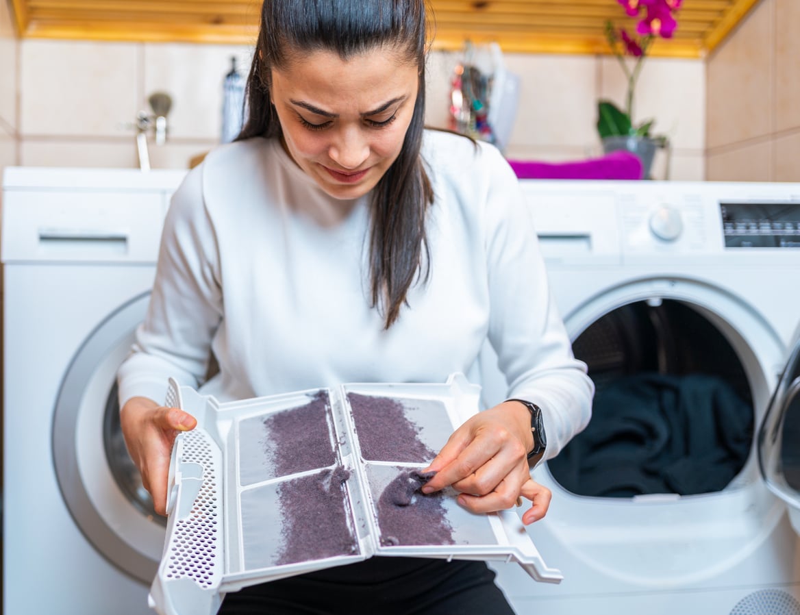 A woman holding a dryer lint trap covered in a thick layer of dust in front of an open clothes dryer.