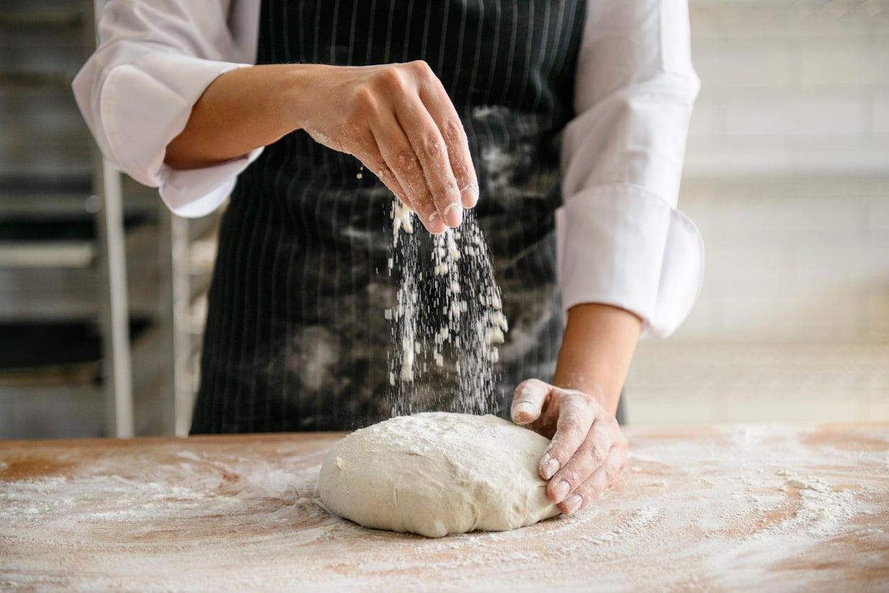 A chef in a white coat and black apron sprinkles flour over a mound of dough on a wooden surface.