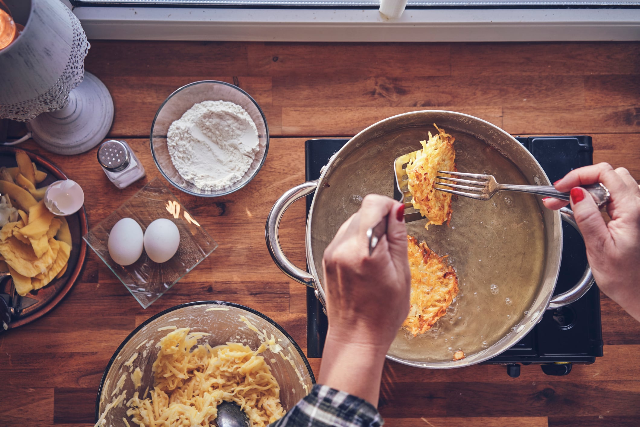 A person uses a spatula and fork to fry potato pancakes, a popular breakfast food, in a pan of oil, with bowls of shredded potatoes, flour, and eggs on the wooden table nearby.
