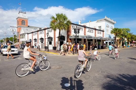 People ride bicycles past Sloppy Joe’s Bar on a sunny day in Key West, Florida. A group stands outside the bar, palm trees line the street, and historic buildings are visible in the background.