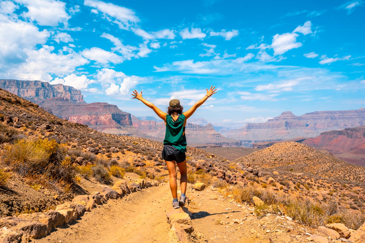 A hiker standing on a dirt trail with arms raised, overlooking a vast canyon under a bright blue sky.