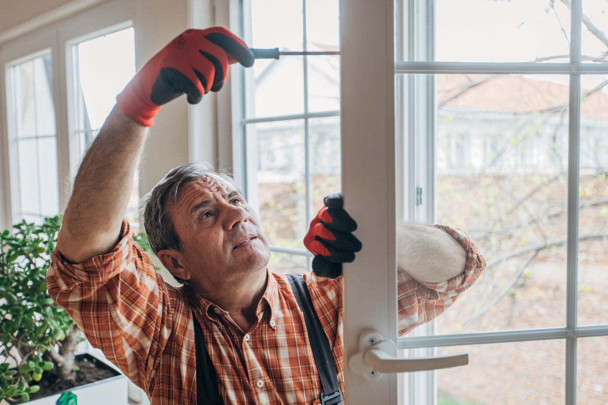 A man in a plaid shirt and work gloves uses a screwdriver to adjust the hinges of a window.