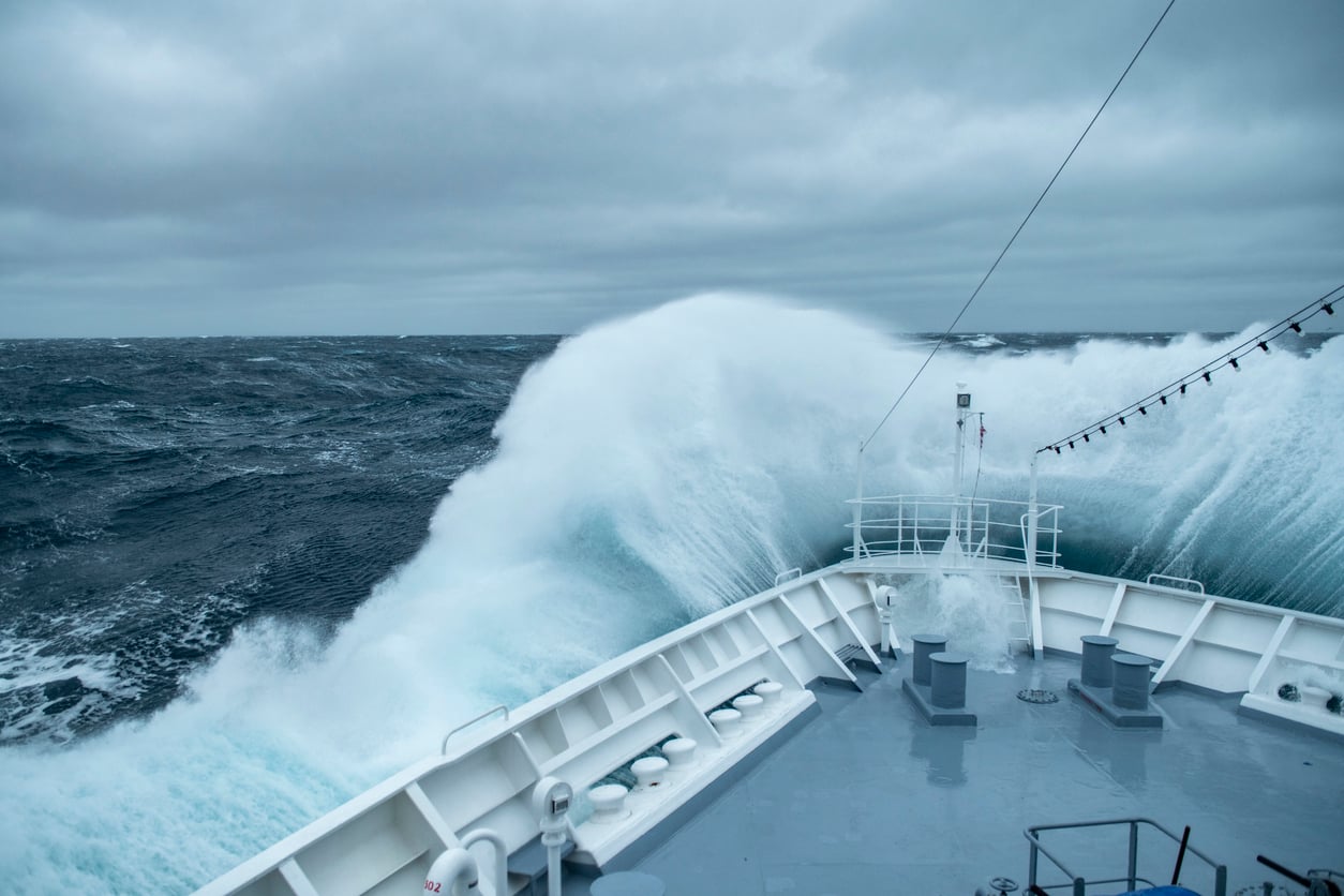 A large wave crashes over the bow of a ship in rough, grey seas under a cloudy sky.