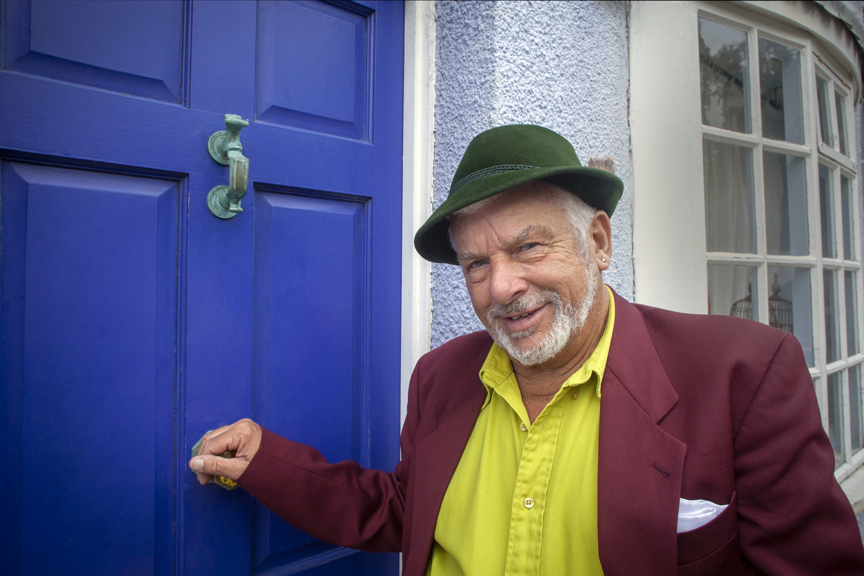 A smiling man wearing a dark green hat, a lime green shirt, and a maroon blazer reaches for the brass handle on a bright blue door.