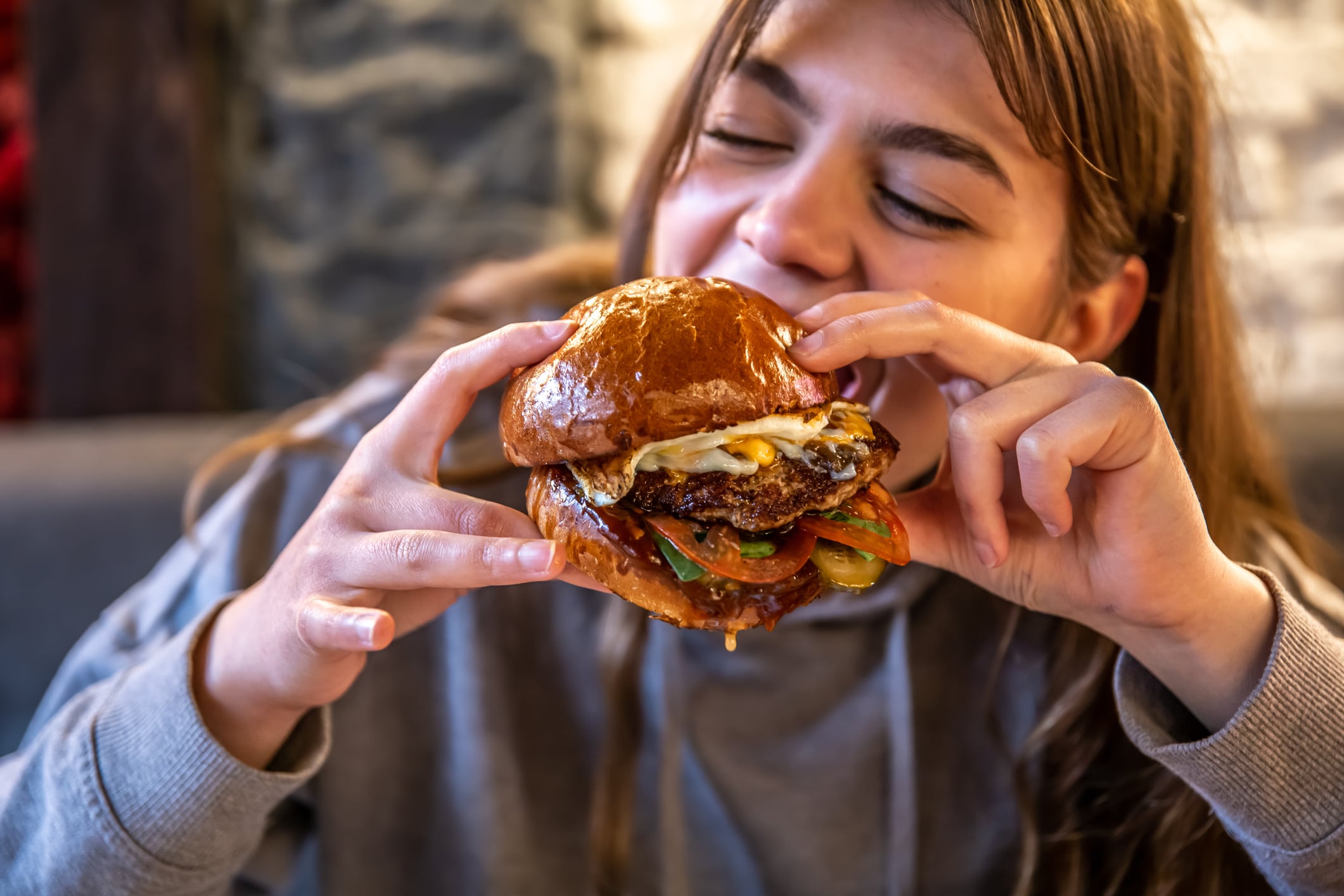 A person with their eyes closed in anticipation holds a breakfast burger topped with a fried egg and vegetables.