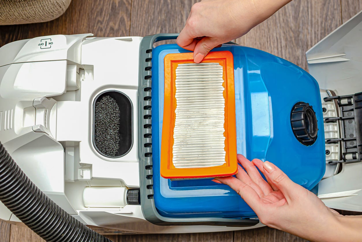 Hands placing a rectangular, orange-framed pleated vacuum filter into a blue and white vacuum cleaner.