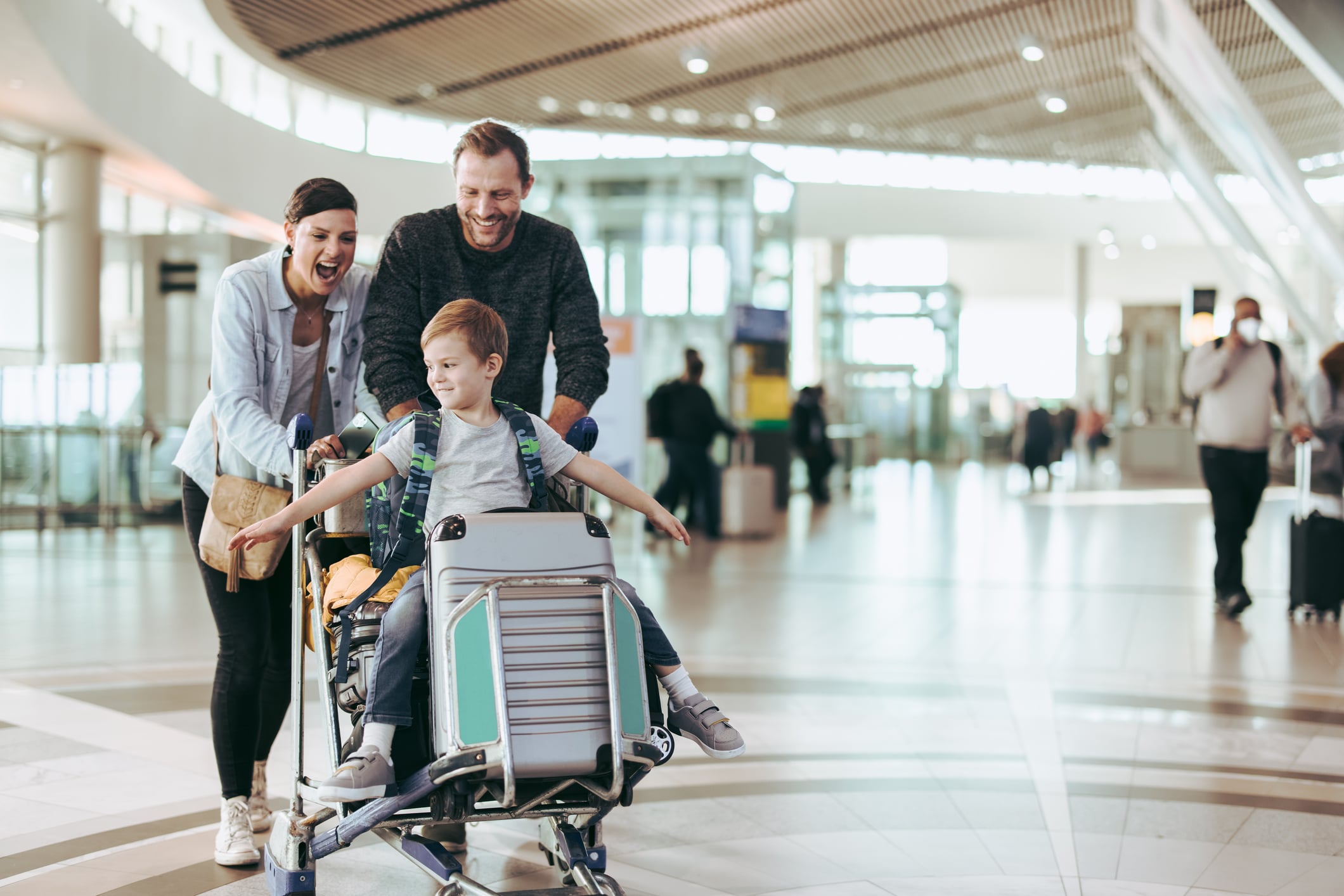 A young boy with arms outstretched sits on a suitcase atop a luggage cart being pushed by his smiling parents through an airport terminal.