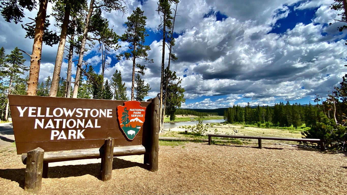 A large wooden Yellowstone National Park sign stands by a scenic area with trees, a river, and a blue sky with clouds.