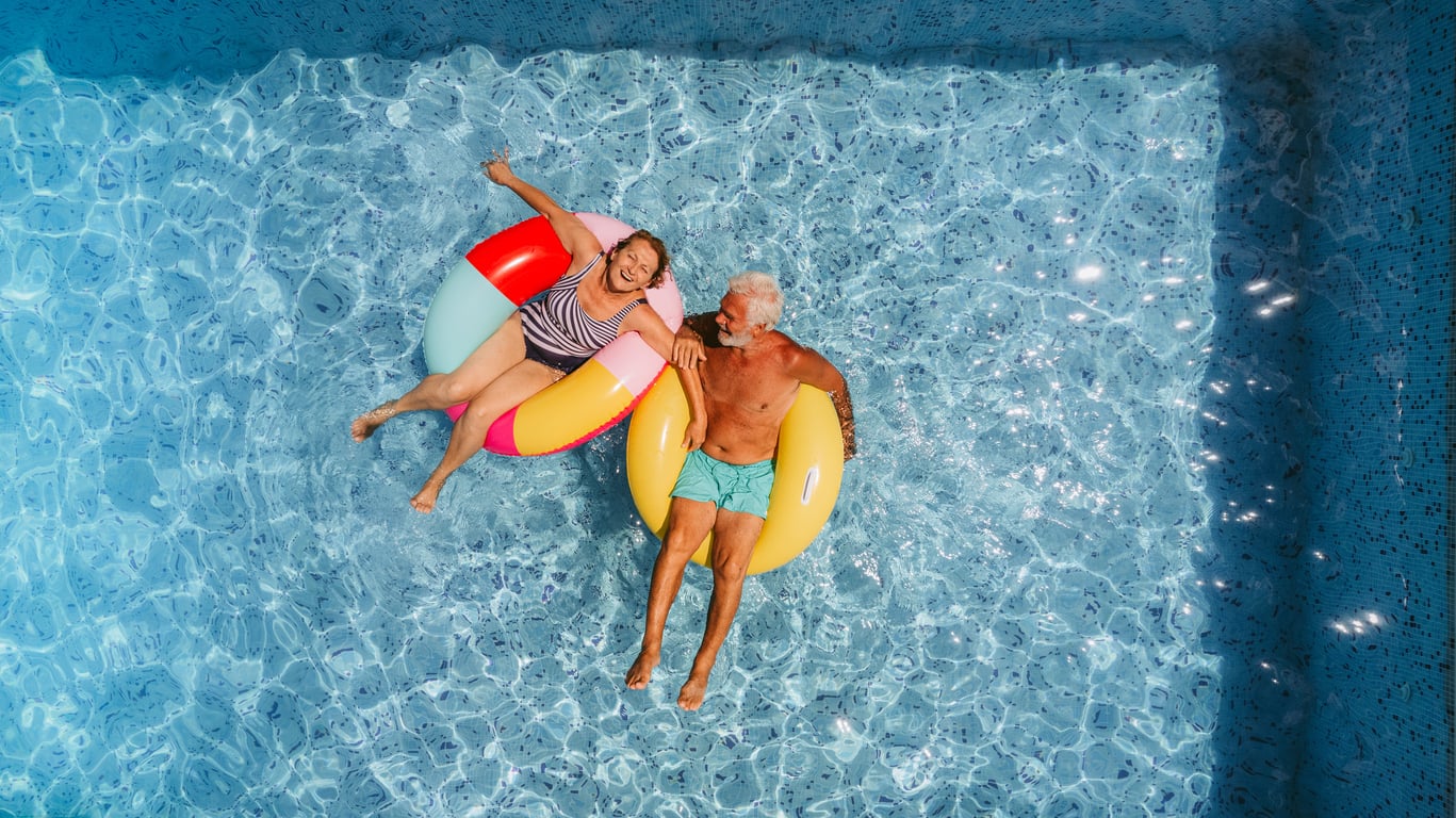 An older couple floating on colorful inner tubes in a swimming pool.