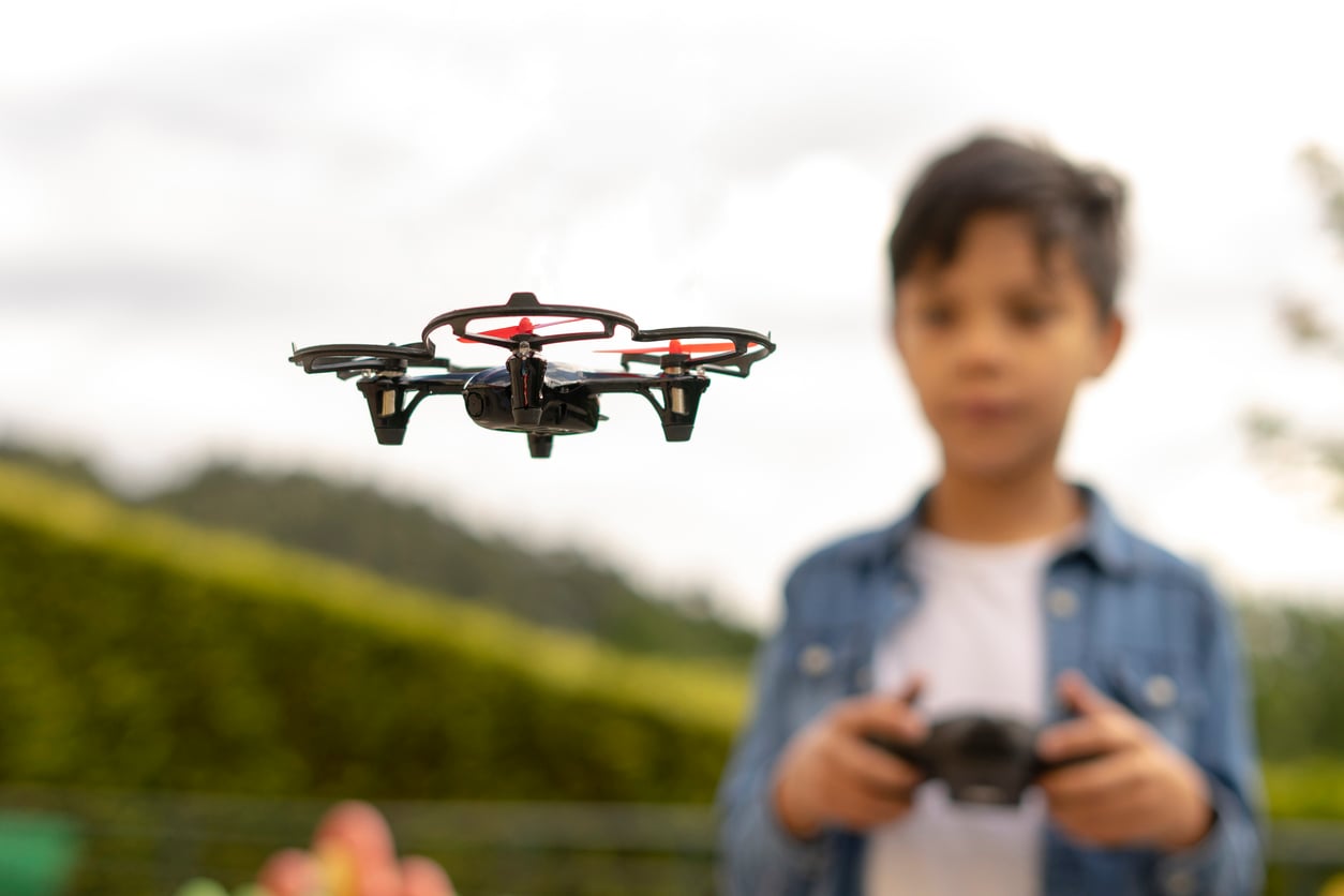 A small drone hovering in mid-air as a young boy controls it with a remote.