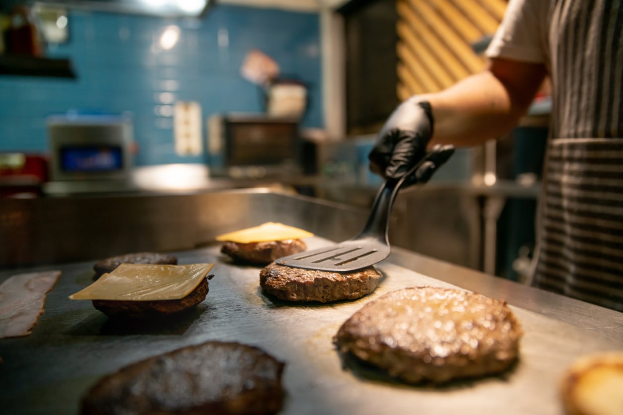 A gloved hand uses a spatula to press down on a burger patty cooking on a flat-top grill alongside other patties topped with melted cheese.