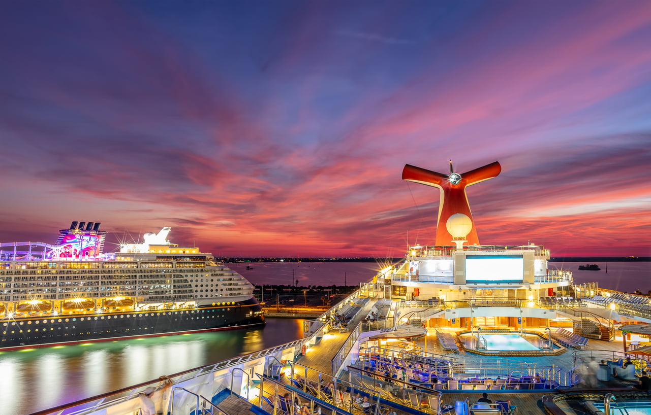 Two large cruise ships docked side-by-side at sunset, with the Carnival Liberty and its iconic red funnel providing a scenic backdrop for cruise entertainment in the foreground.