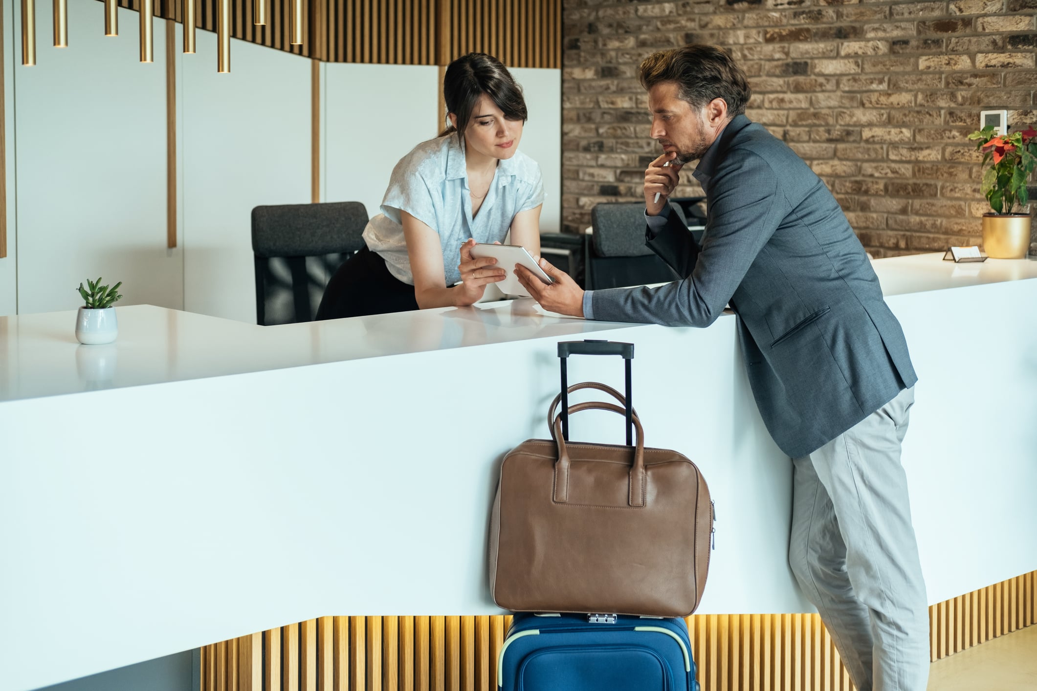 A hotel receptionist and a man in a business suit reviewing information on a tablet at a front desk, with a suitcase sitting on the floor nearby.