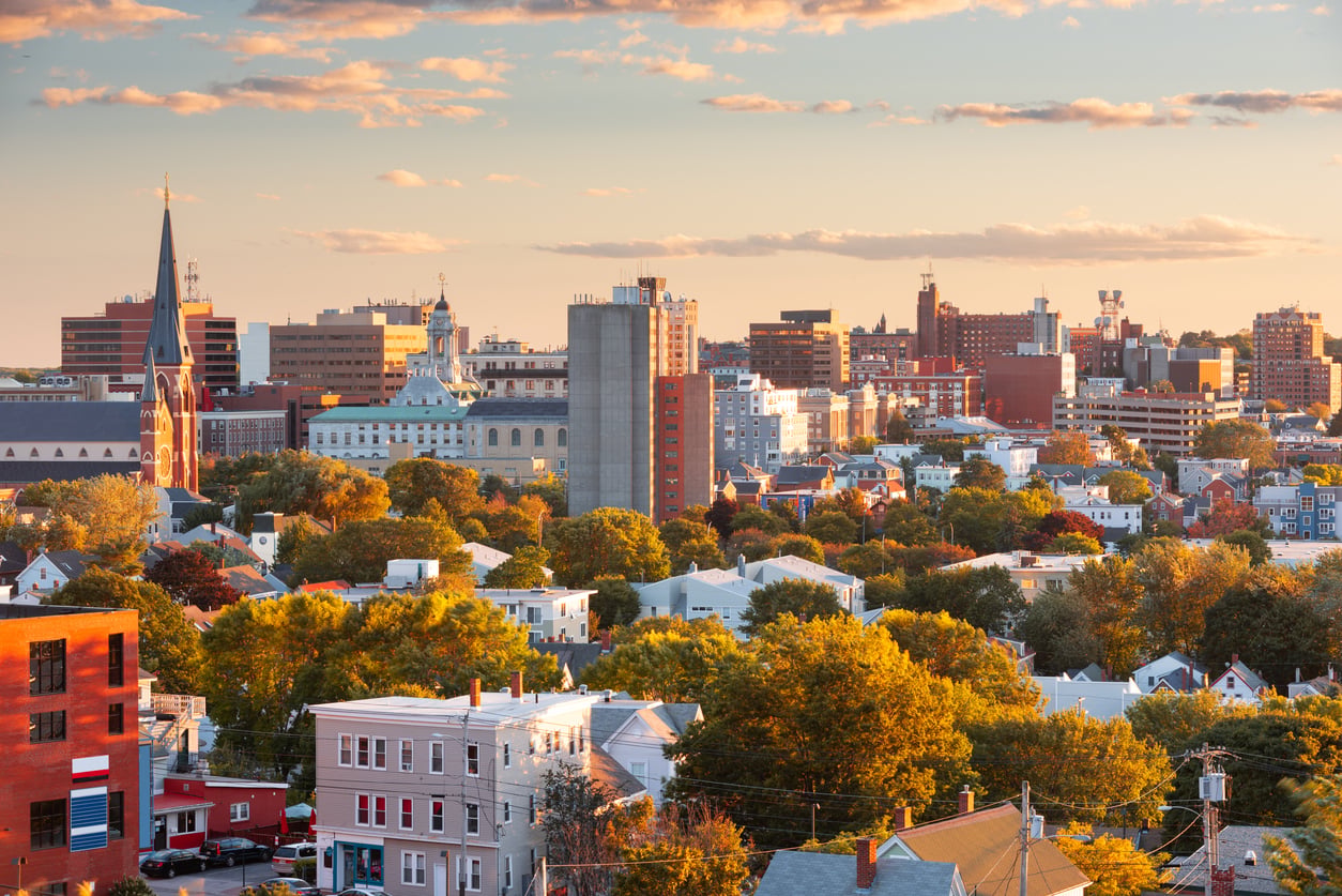 City skyline at sunset with a mix of modern buildings, a church steeple, and tree-filled neighborhoods. Warm sunlight casts a golden hue over one of those American Destinations That Feel Bigger than you’d expect.