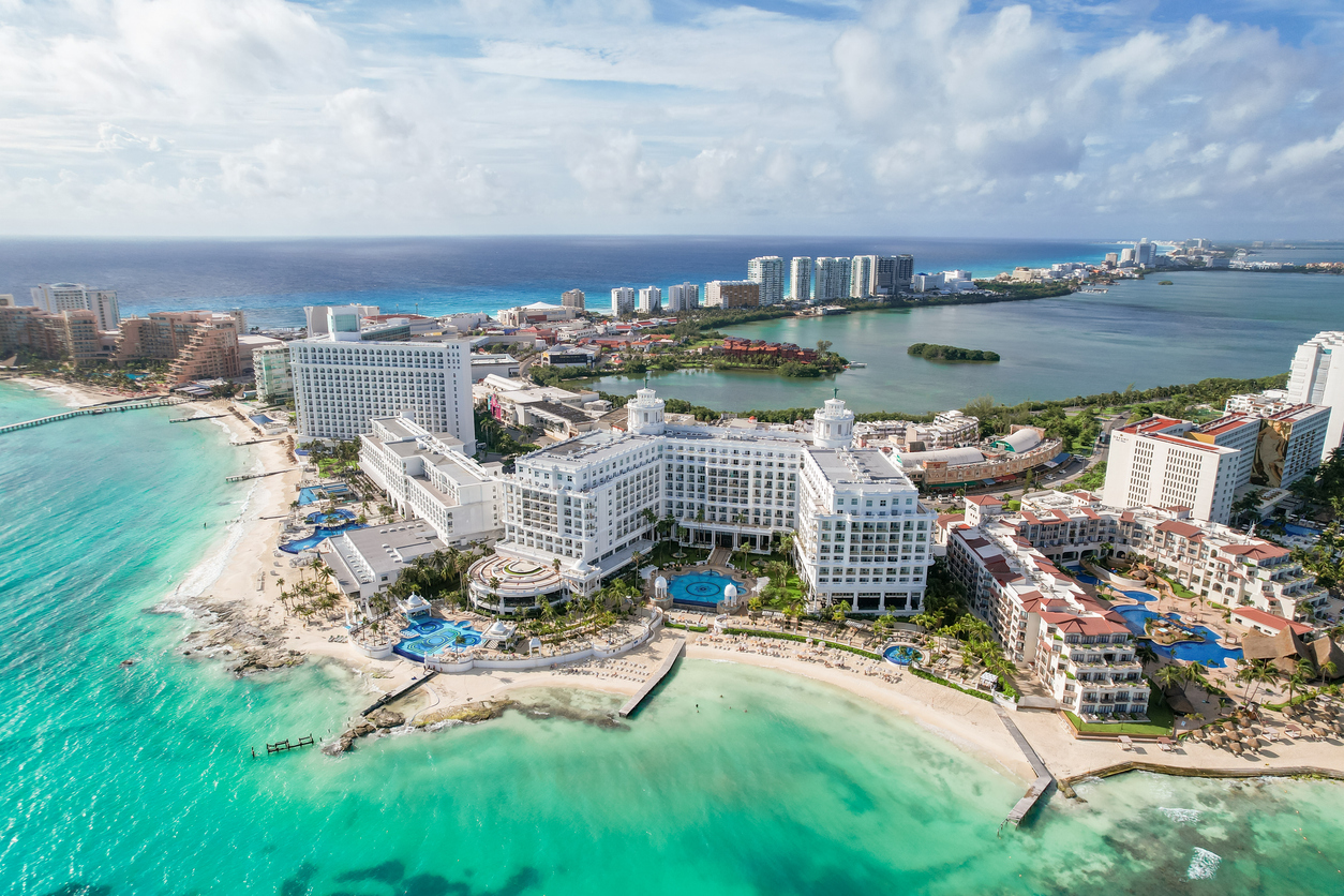 Aerial view of a luxurious white resort complex situated on a beach with turquoise water, bordering a lagoon and a distant city skyline.