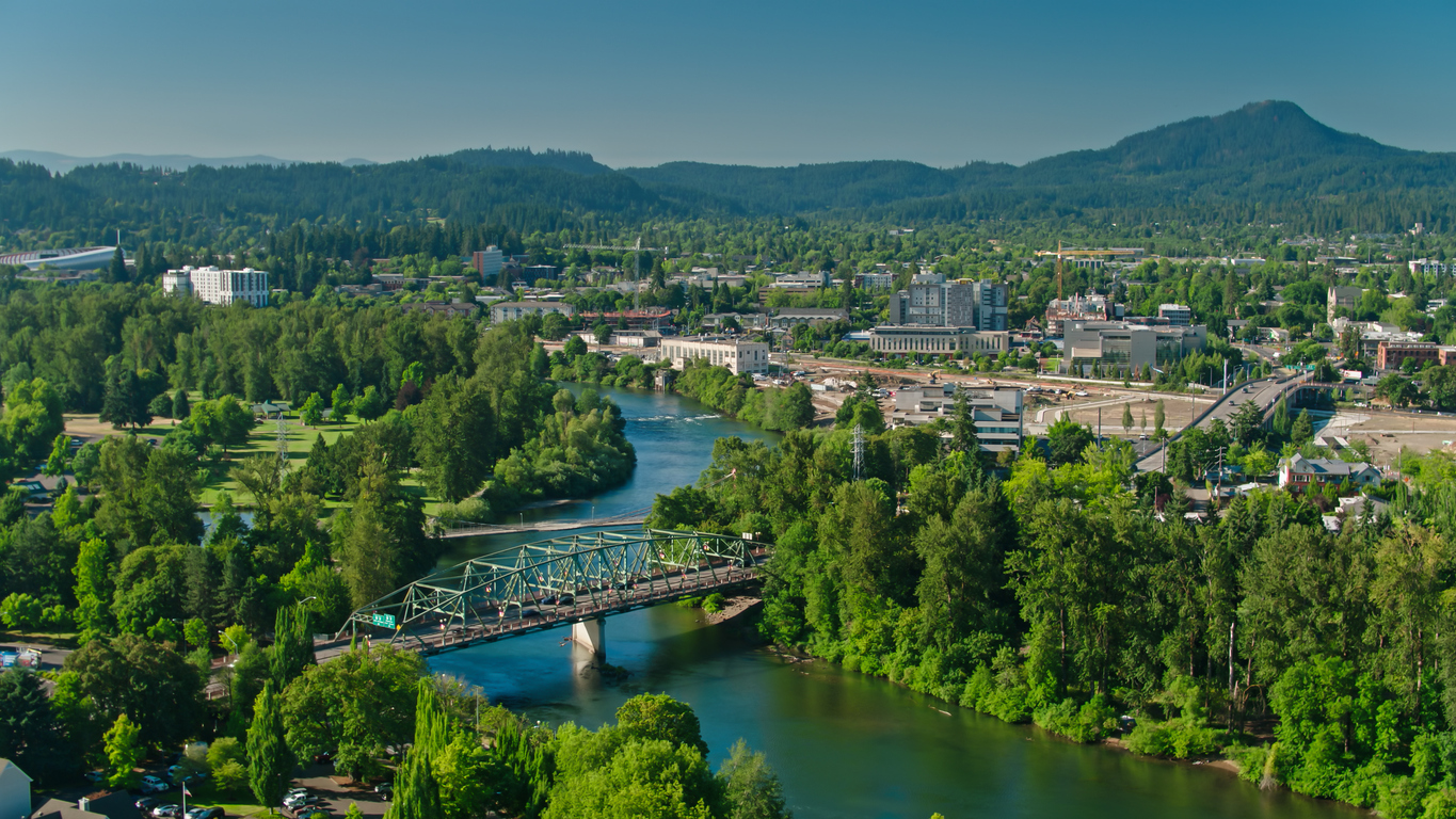Aerial view of a green truss bridge spanning a river winding through a town surrounded by dense forests and mountains.