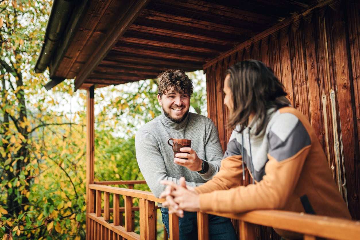 Two men talk while leaning on a wooden porch railing, one holding a mug.