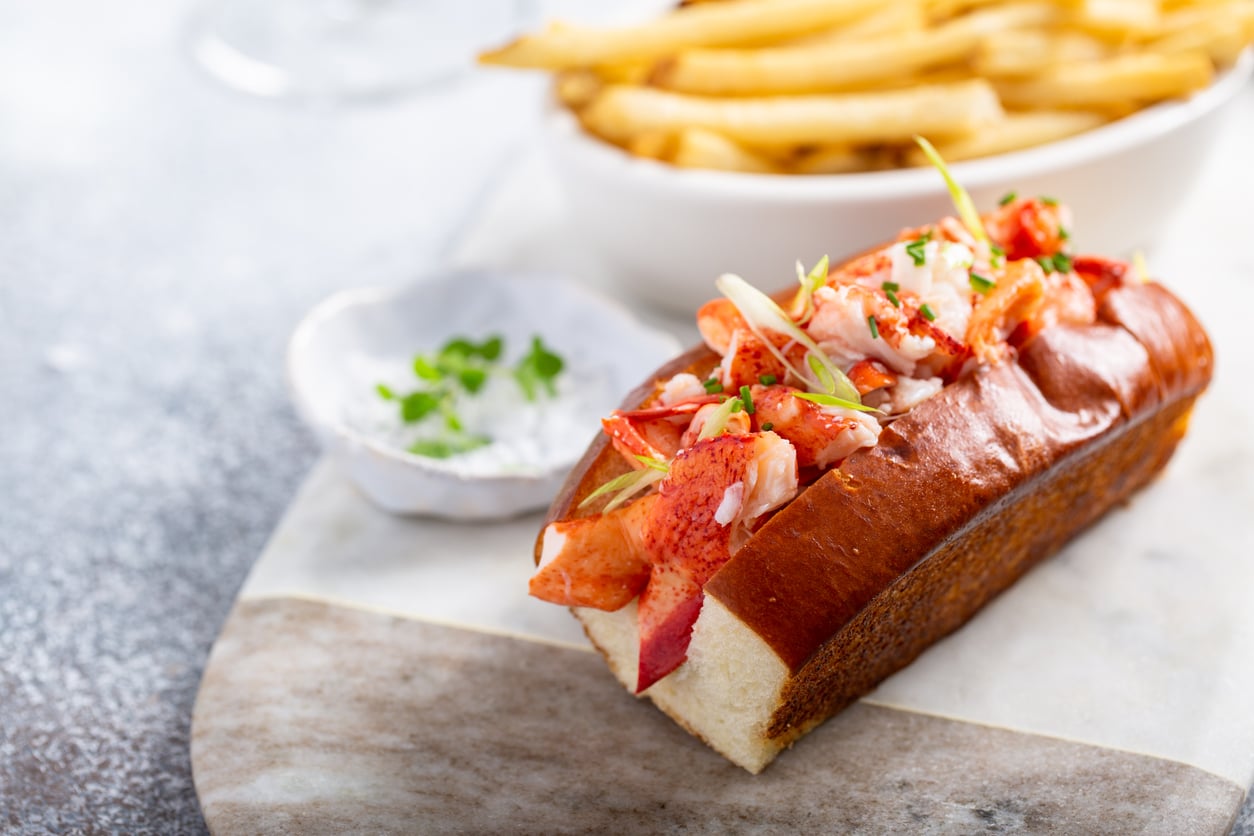 A lobster roll in a toasted bun, served with a side of french fries and a small bowl of dipping sauce, a good food quality.