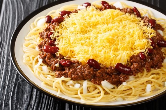 Spaghetti topped with chili, shredded cheddar cheese, kidney beans, and diced white onions on a plate.