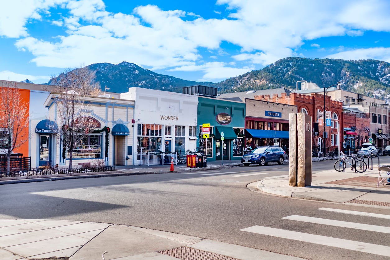 A street view of a downtown block with various shops including Old Tibet, Wonder, and Trident, with mountains visible in the background under a blue sky.