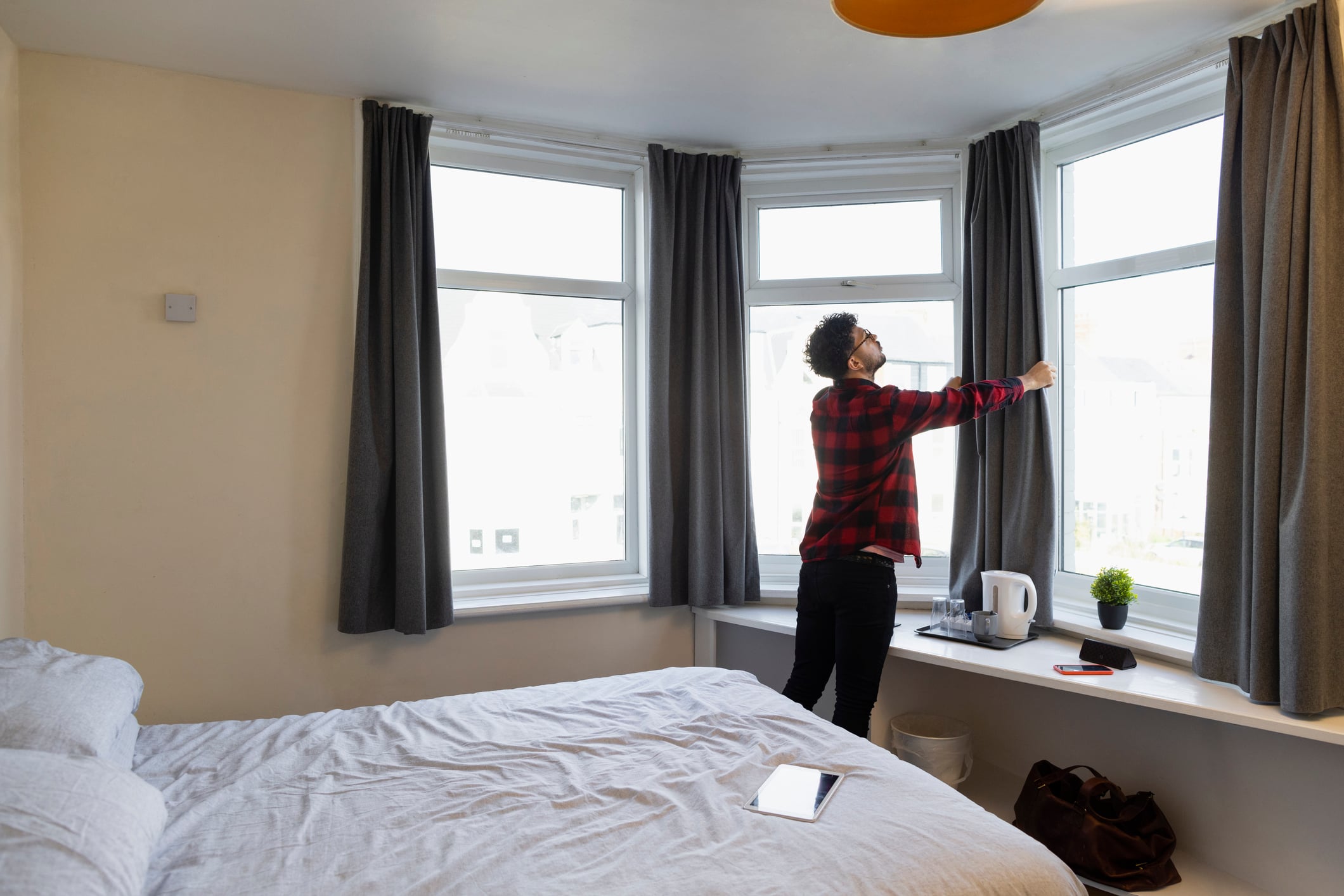 Things you should do when staying in a hotel: A person in a red plaid shirt stands by a bedroom window, pulling the curtain closed.